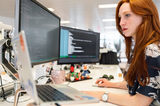 A red-haired white woman focuses on a desk with two monitor screens and one laptop in front of her.