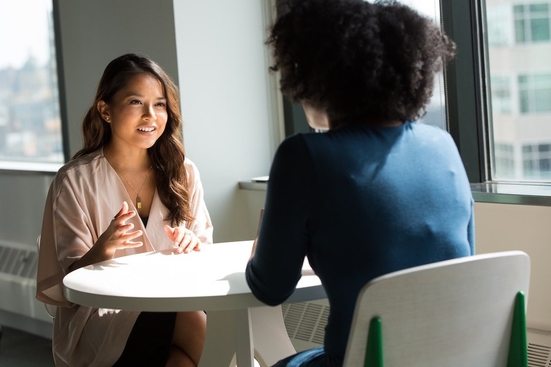 Two women wearing professional clothing sitting at a table having a conversation. 