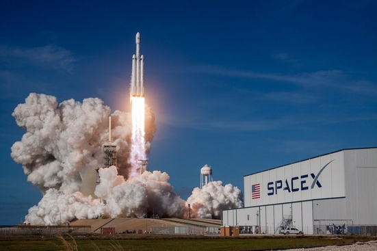 A SpaceX rocket launching beside a building with an American flag and the SpaceX logo