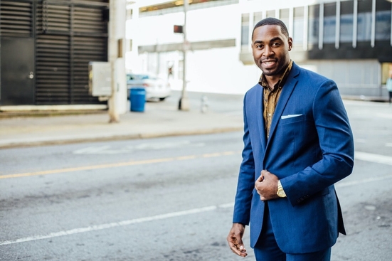 A Black man in a navy blue suit walking down a city street and smiling into the camera