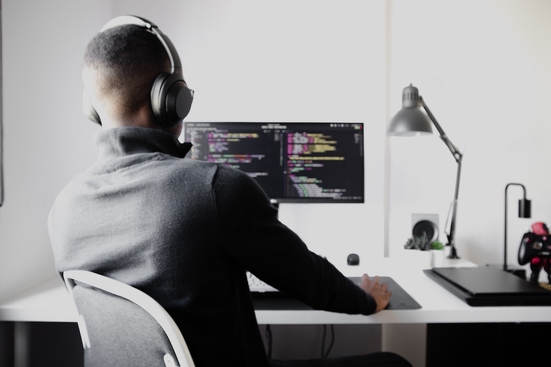 A Black person with close-cropped hair wearing headphones sits at a desk with code across the monitor screen