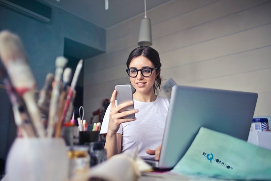 A white woman with glasses sitting in front of a laptop looking at a phone